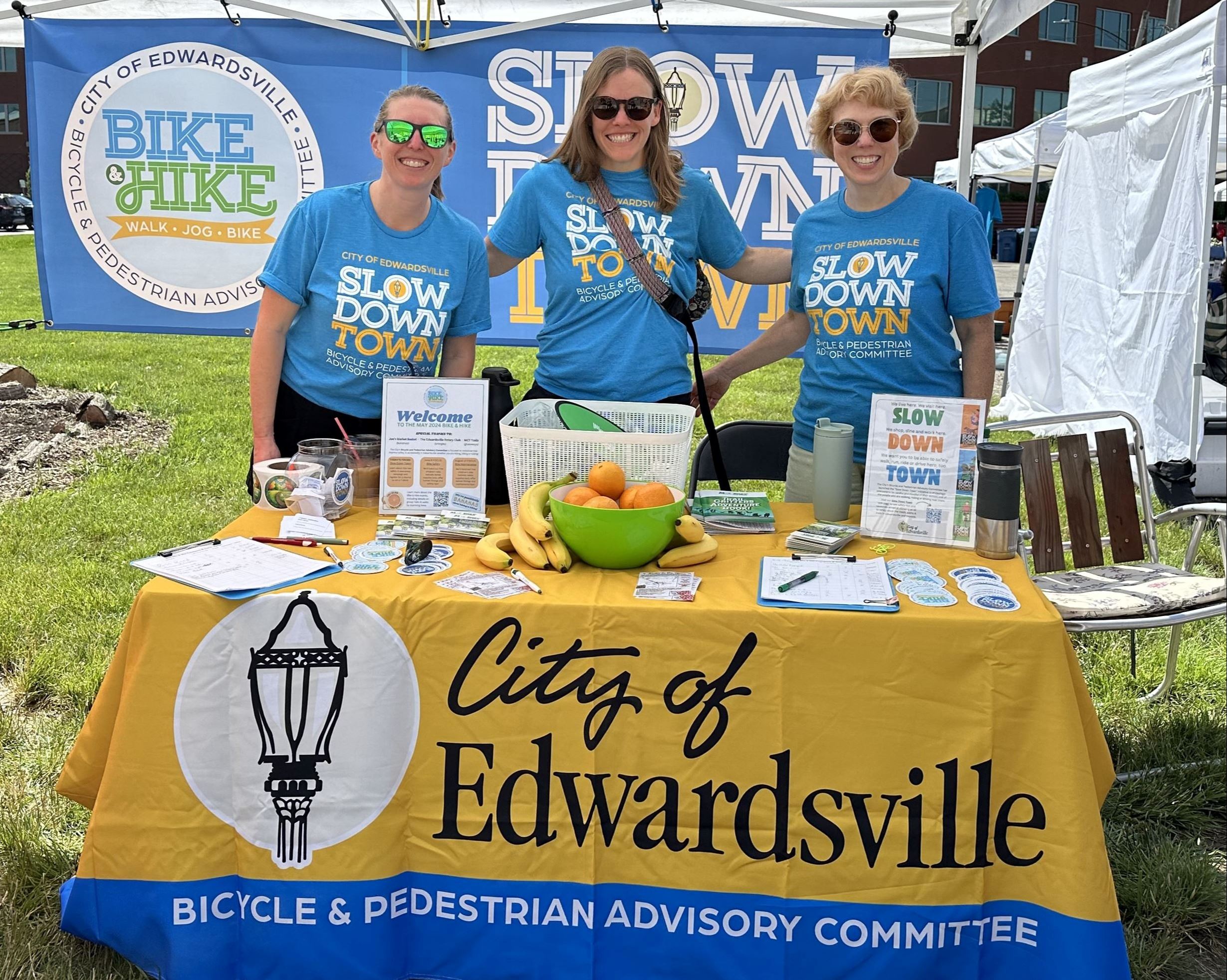 Three people in matching blue shirts stand behind a table in a tent outside