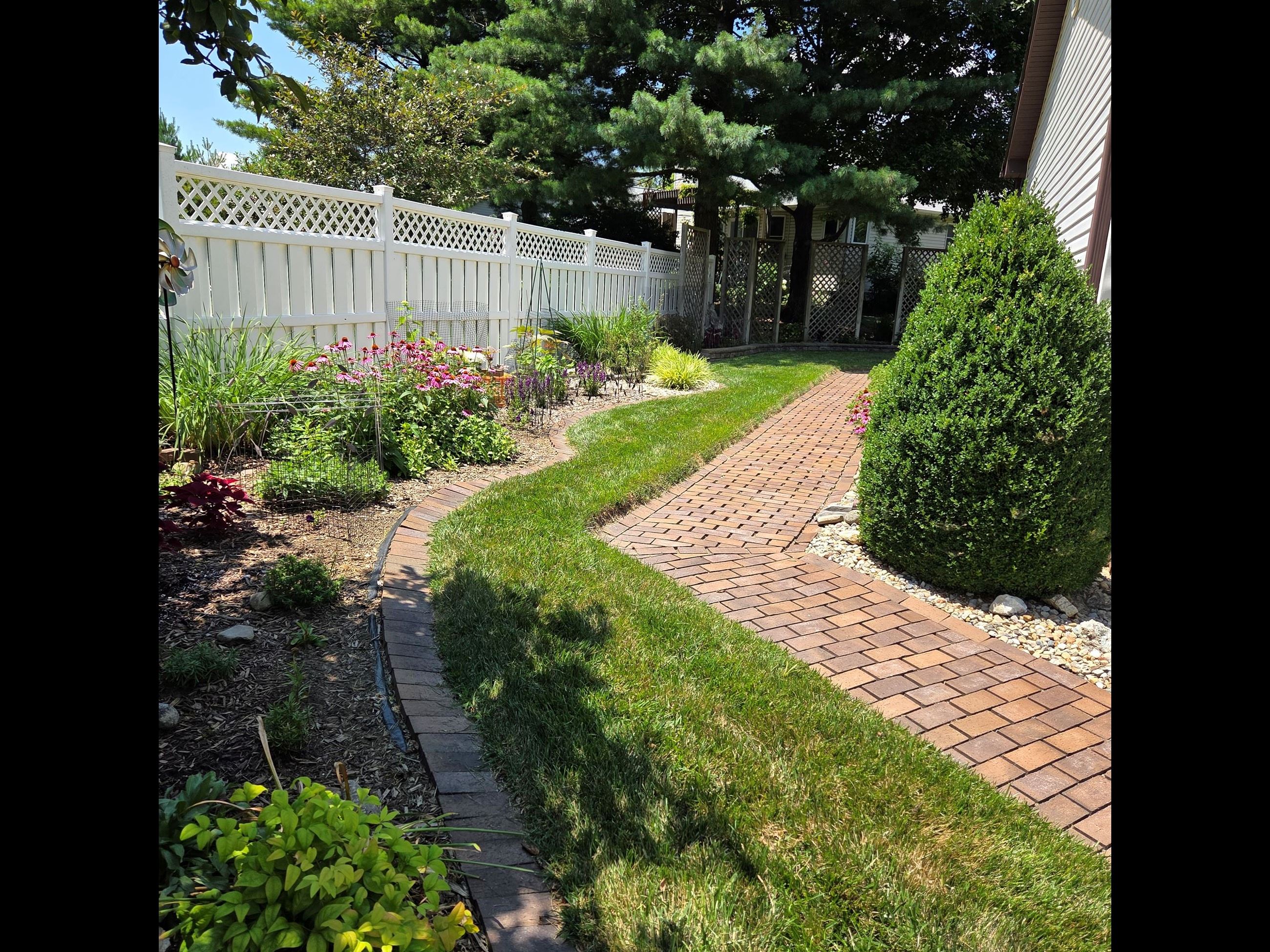 Image shows flowers, shrubs, a fence and walkway along a home