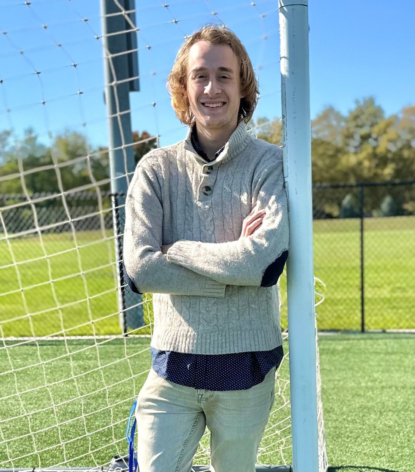 A tall thin man in a sweater stands next to a soccer net in a grassy field